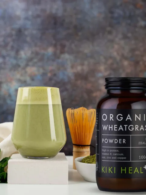 Organic wheatgrass powder jar, glass of green drink, whisk, and mint leaves on a white surface.