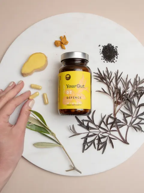 YourGut 02 Defence supplement bottle with herbs, ginger, and capsules on a round white surface.