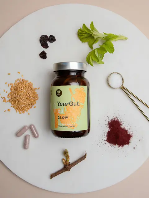 YourGut Glow supplement jar with herbs, seeds, capsules, and a spoon of salt on a round board.