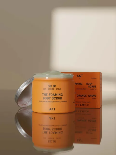 Orange AKT foaming body scrub jar and box on reflective surface.