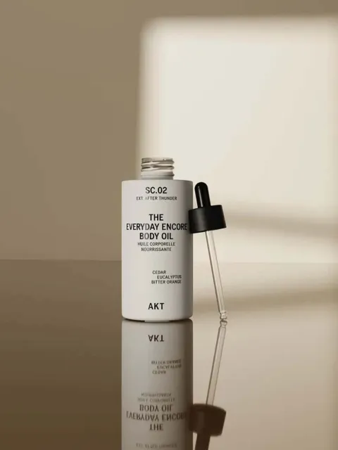 Open bottle of AKT Everyday Encore Body Oil with dropper on reflective surface.
