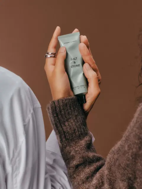 Hands holding a Salt & Stone cream tube against a brown background.