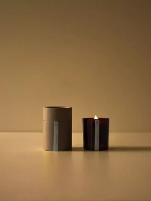 Lit candle in a dark glass holder next to a brown cylindrical box on a beige surface.