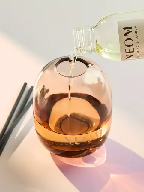 Diffuser oil being poured into a NEOM glass diffuser with reeds beside it.