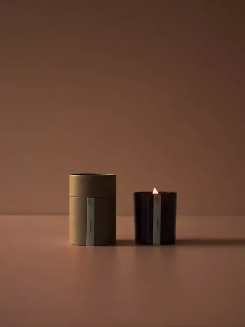 Lit candle in a dark glass holder next to a brown cylindrical box on a brown surface.