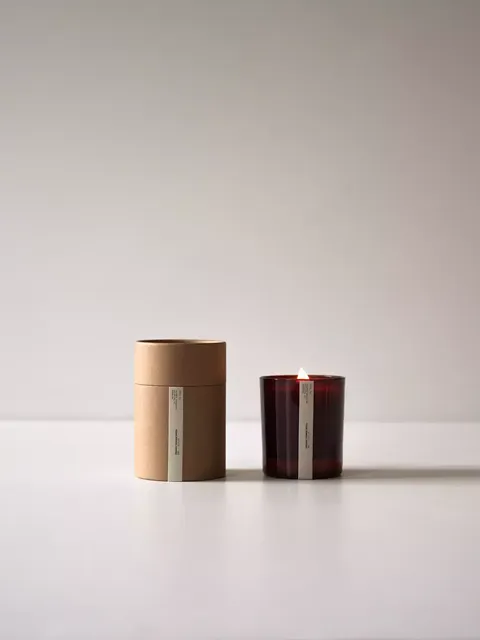 Brown candle box and lit red glass candle on white surface.