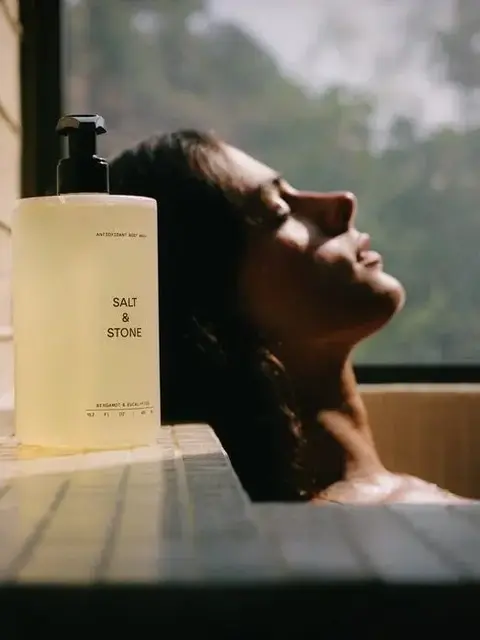 Bottle of Salt & Stone body wash on tiled ledge, person relaxing in bath by window.