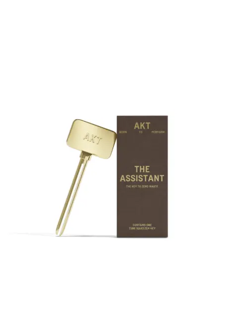 Gold tube squeezer key with "AKT" branding next to a brown box labelled "The Assistant".