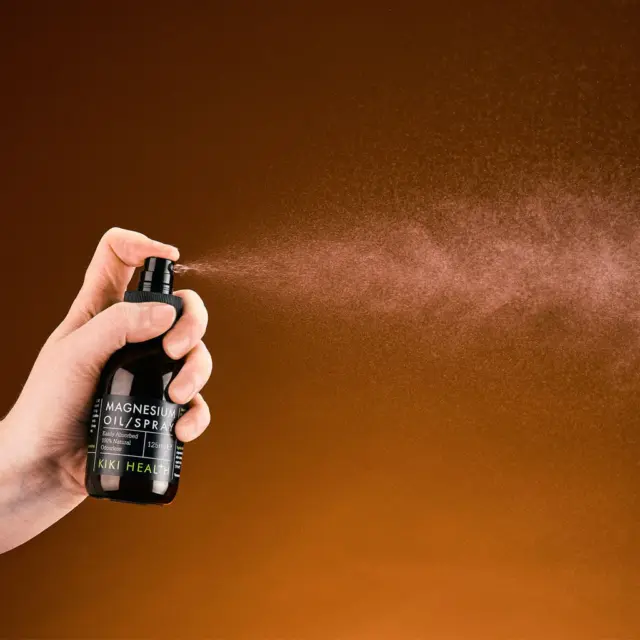 Hand spraying magnesium oil from a black bottle against a brown background.