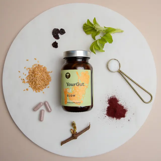 YourGut Glow supplement jar with herbs, seeds, capsules, and a spoon of salt on a round board.