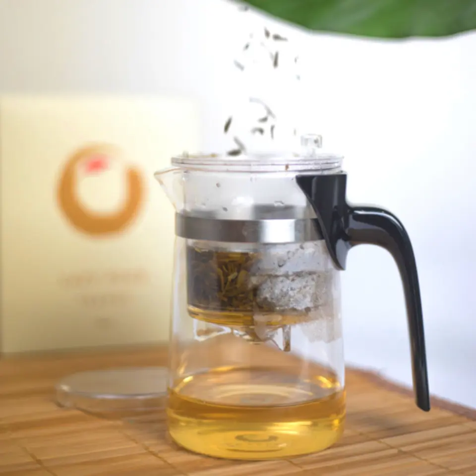 Glass teapot with infuser brewing tea, on a bamboo mat. Box with circular logo in background.