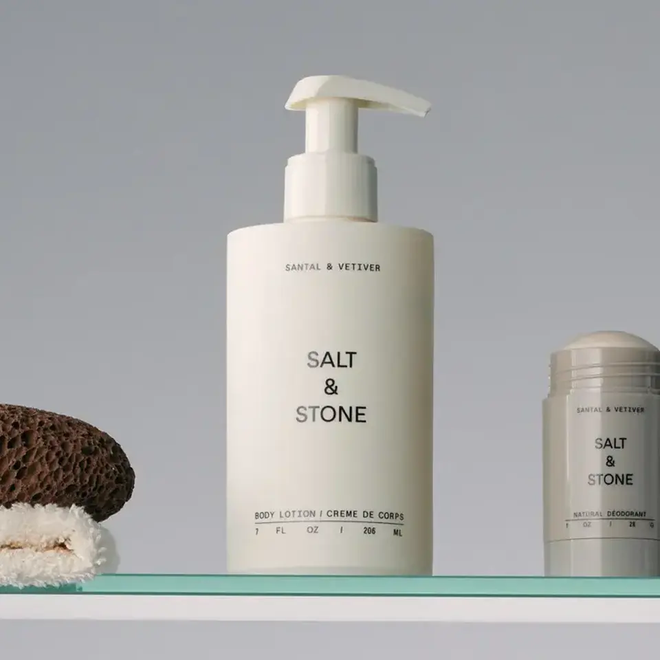 Salt & Stone body lotion and deodorant on a glass shelf with a pumice stone and towel.