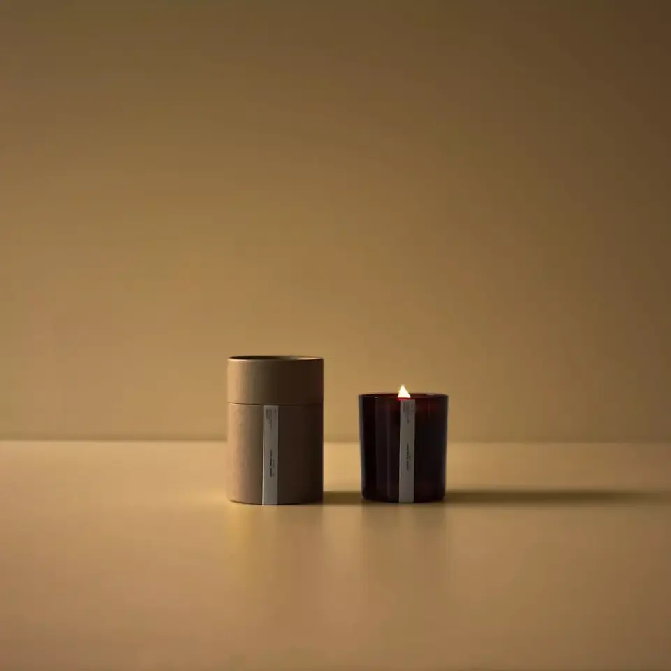 Lit candle in a dark glass holder next to a brown cylindrical box on a beige surface.