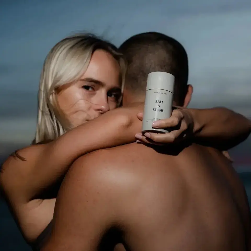 Woman holding Salt & Stone product, embracing a man at sunset by the sea.