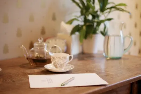 Teapot, teacup, paper, and pen on wooden table with plant and water jug in background.