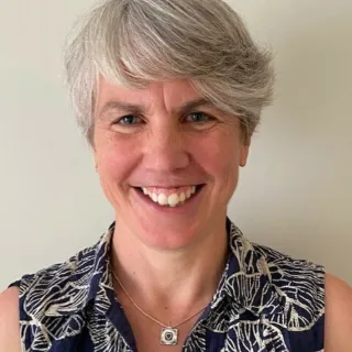 Smiling person with short grey hair, wearing a patterned sleeveless top and necklace.