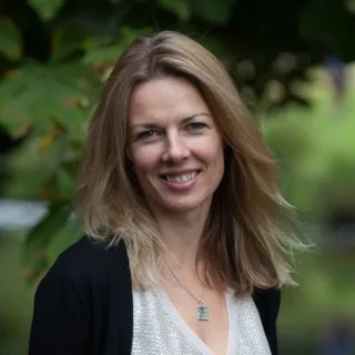 Woman with long blonde hair smiling outdoors, wearing a black cardigan and silver necklace.