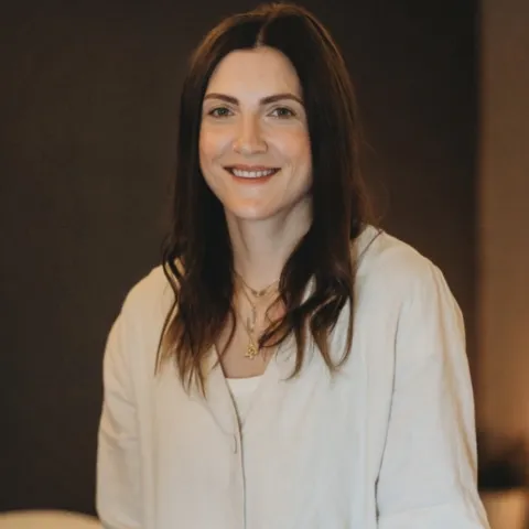 Woman with long hair in a light-coloured shirt, smiling indoors.