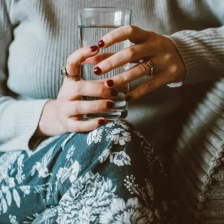 Person holding a glass of water, wearing a grey jumper and floral trousers.