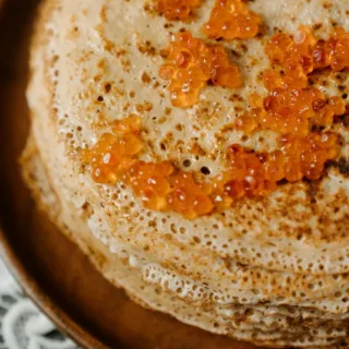 Stack of pancakes topped with red caviar on a wooden plate, lace tablecloth underneath.