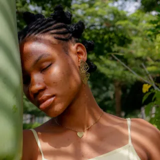 Woman with braided hair leans against a tree, wearing a heart necklace and leaf-shaped earring.