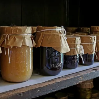Jars with paper covers on a wooden shelf, containing various preserves and pickles.