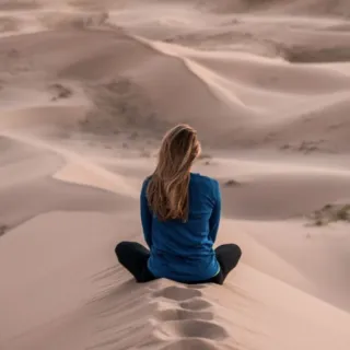 Person sitting on sand dune, facing vast desert landscape.