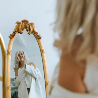 Woman in white outfit smiling at her reflection in an ornate mirror.