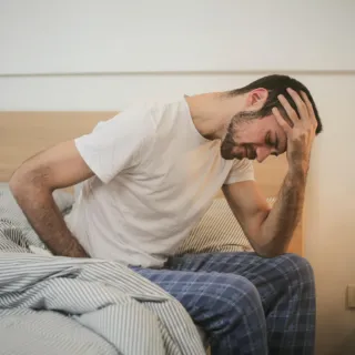 Man in pyjamas sitting on bed, holding head with hand, appearing unwell.
