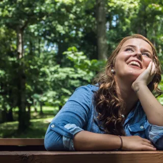 Woman in denim shirt smiling outdoors in a wooded area.