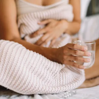 Person sitting on bed holding a glass of water, wrapped in a knitted blanket.