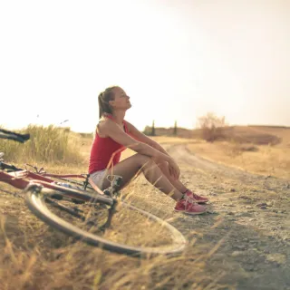 Woman in sportswear sitting on a dirt path beside a bicycle in a rural landscape.