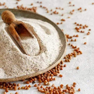 Buckwheat flour with a wooden scoop on a plate, surrounded by buckwheat grains.