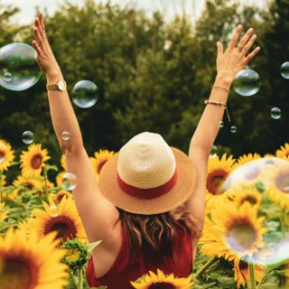 Woman in a sunflower field with arms raised, wearing a straw hat. Bubbles float around her.