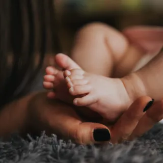 Baby's feet resting on an adult's hand with black nail polish, on a soft grey surface.
