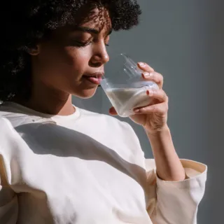 Person drinking from a glass of milk, wearing a light-coloured top, in natural light.