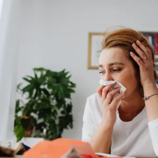 Woman holding tissue to nose, sitting at table with papers, plant in background.