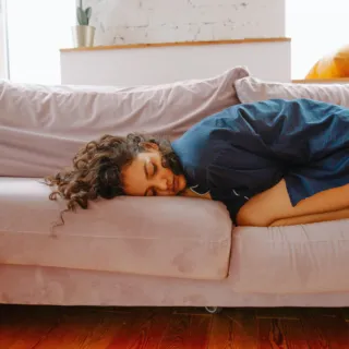 Person sleeping on a light purple sofa, wearing a blue dress. Yellow cushion in the background.