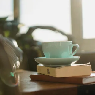 Teal cup on saucer atop two books on a wooden table in a sunlit room.