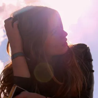 Woman with long hair in sunlight, holding phone, sky and clouds in background.