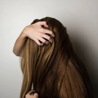 Person with long brown hair, hand touching scalp against a plain wall.