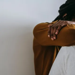 Person in brown jumper resting head on arms, sitting against a white wall.