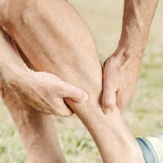 Person holding their calf muscle outdoors, wearing blue shorts and trainers.