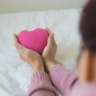 Hands holding a pink heart-shaped cushion on a white bed.