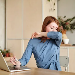 Woman in blue top sneezing into elbow at desk with laptop in home office setting.