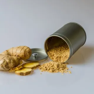 Ginger root and slices next to a tipped metal tin of ginger powder on a white surface.