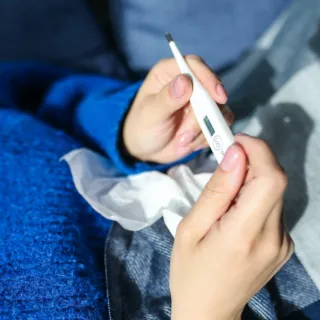 Person holding a digital thermometer and tissue, wearing a blue jumper.