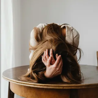 Person with head down on table, hands in hair, seated in a wooden chair.