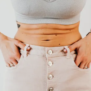 Person in grey crop top and beige buttoned skirt, hands in pockets, showing midriff tattoos.
