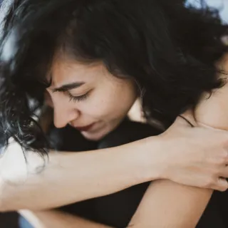 Person with dark hair hugging knees, wearing a black top, indoors.